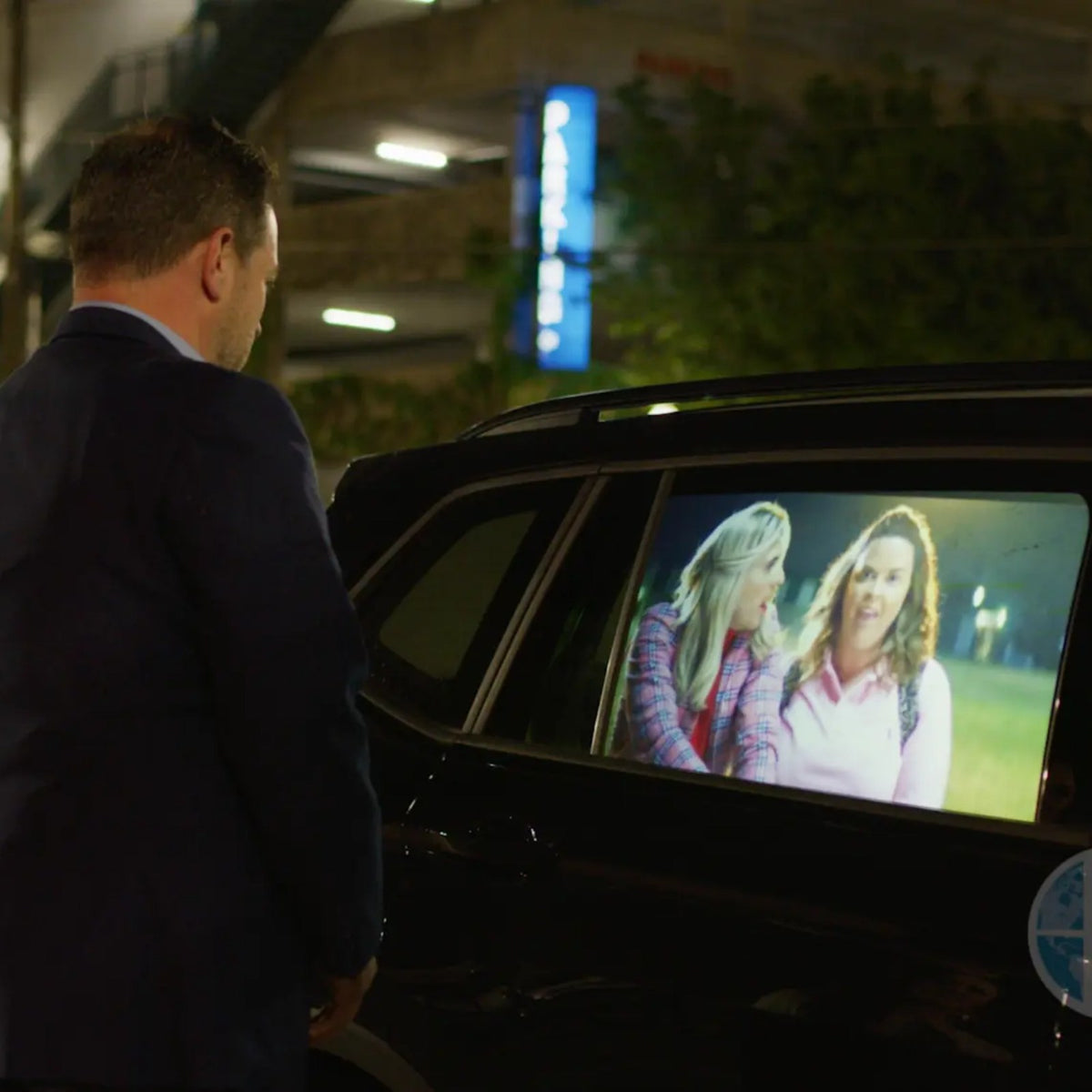 HoloGlass rear side door window display playing a video on a parked SUV at night, with a man watching